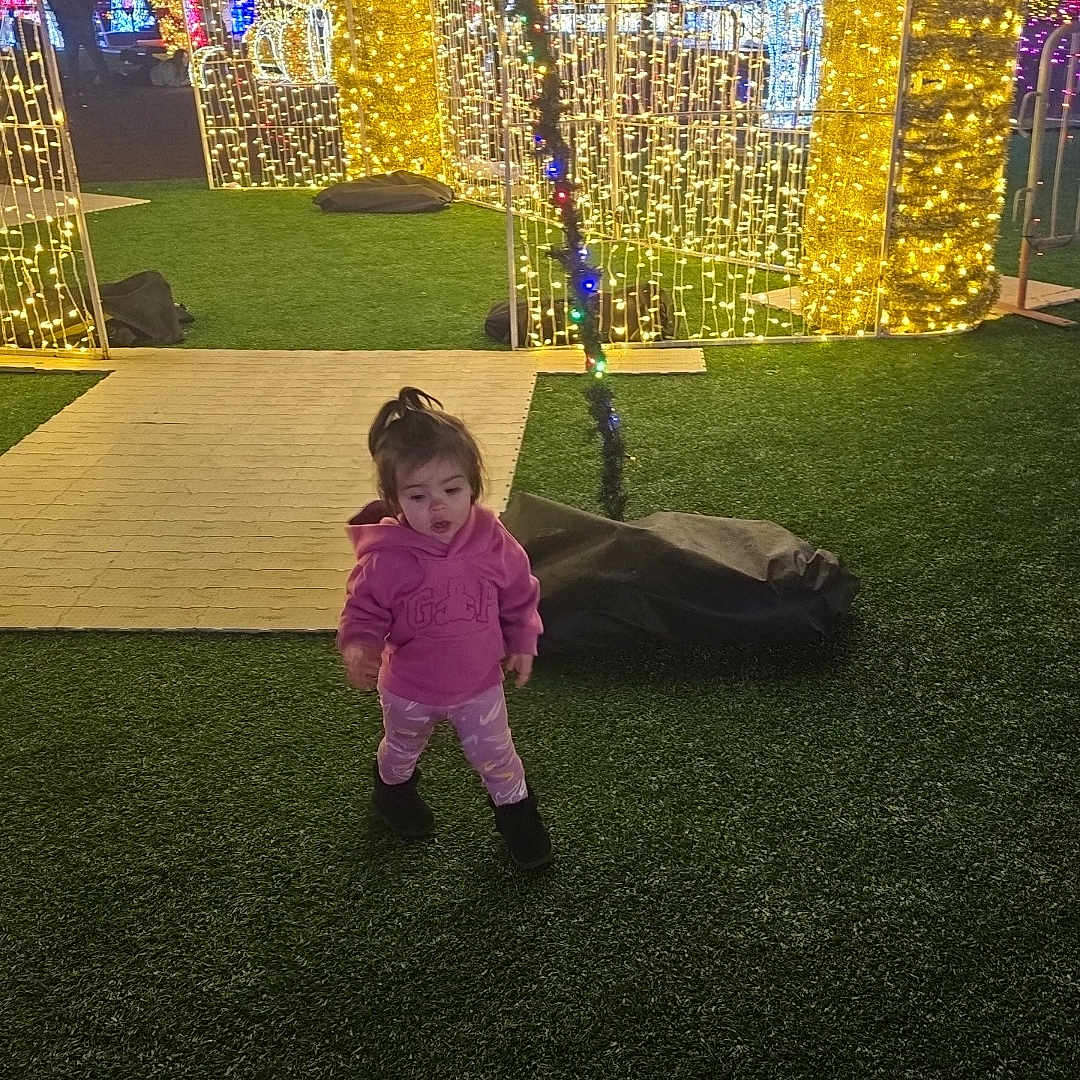 Evelyn is registered to the contest to win money with this photo: arch, celebration, child, colorful_lights, decoration, festive, festivity, fun, grass, holiday_lights, illumination, leggings, night, outdoor, person, pink_hoodie, toddler, toy, walking, winter