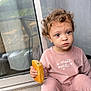 cars, casual, child, curly_hair, curtain, face, floor, food, glass_door, hand, home, outdoor, pastry, pink_clothing, portrait, reflection, serious_expression, sitting, window, young_child