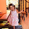 child, toddler, girl, dress, stairs, holding_railing, curly_hair, smiling, indoors, archway, orange_wall, sandals, lighting, stone_steps, iron_railing, playful, portrait, cute, fashion, architecture