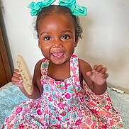 Luna is registered to the contest to win money with this photo: child, toddler, smiling, dress, floral_pattern, bows, hair_accessories, indoor, blanket, happy, cute, person, portrait, buttoned_dress, wooden_object, sitting, bright_eyes, hands, skin, baby
