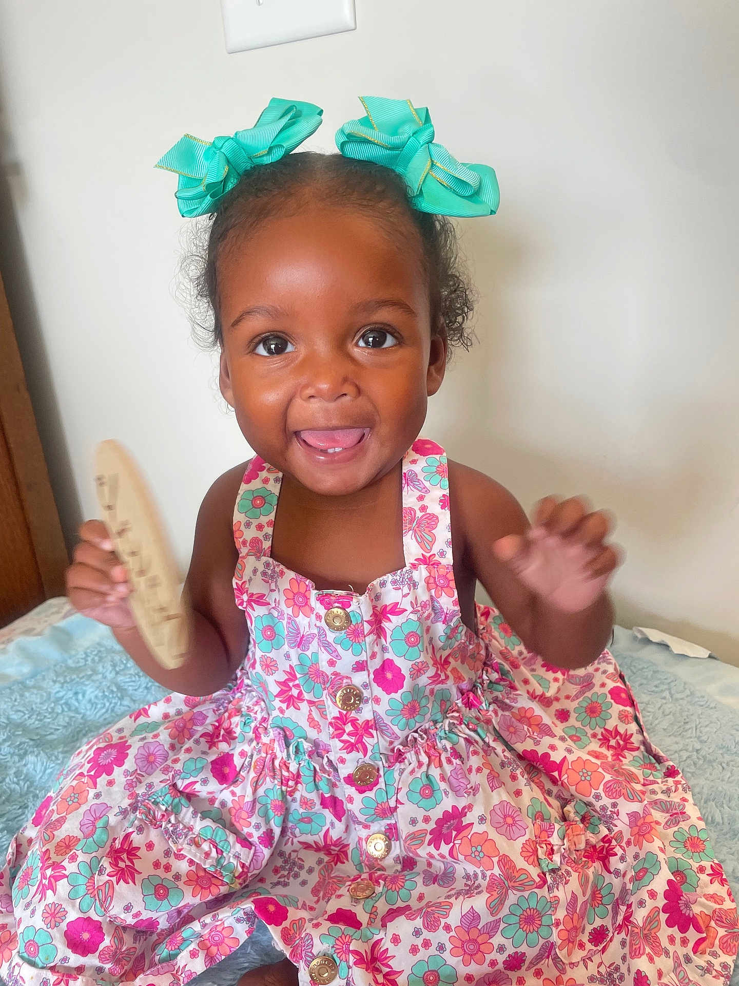 Luna is registered to the contest to win money with this photo: child, toddler, smiling, dress, floral_pattern, bows, hair_accessories, indoor, blanket, happy, cute, person, portrait, buttoned_dress, wooden_object, sitting, bright_eyes, hands, skin, baby