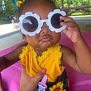 Luna joined the competition — help win amazing prizes! accessory, baby, child, curly_hair, cute, fashion, flower_pattern, hand, outdoor, pavilion, person, pink_nail_polish, playful, portrait, shade, summer, sunflower, sunglasses, toddler, yellow_clothing
