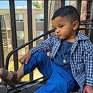 Liam Herrera Zarraga joined the competition — help win amazing prizes! child, toddler, boy, portrait, sitting, chair, outdoor, balcony, patio, blue_clothing, plaid_shirt, pants, sandals, necklace, short_hair, looking_away, face, apartment_building, railing, daylight