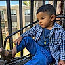child, toddler, boy, portrait, sitting, chair, outdoor, balcony, patio, blue_clothing, plaid_shirt, pants, sandals, necklace, short_hair, looking_away, face, apartment_building, railing, daylight