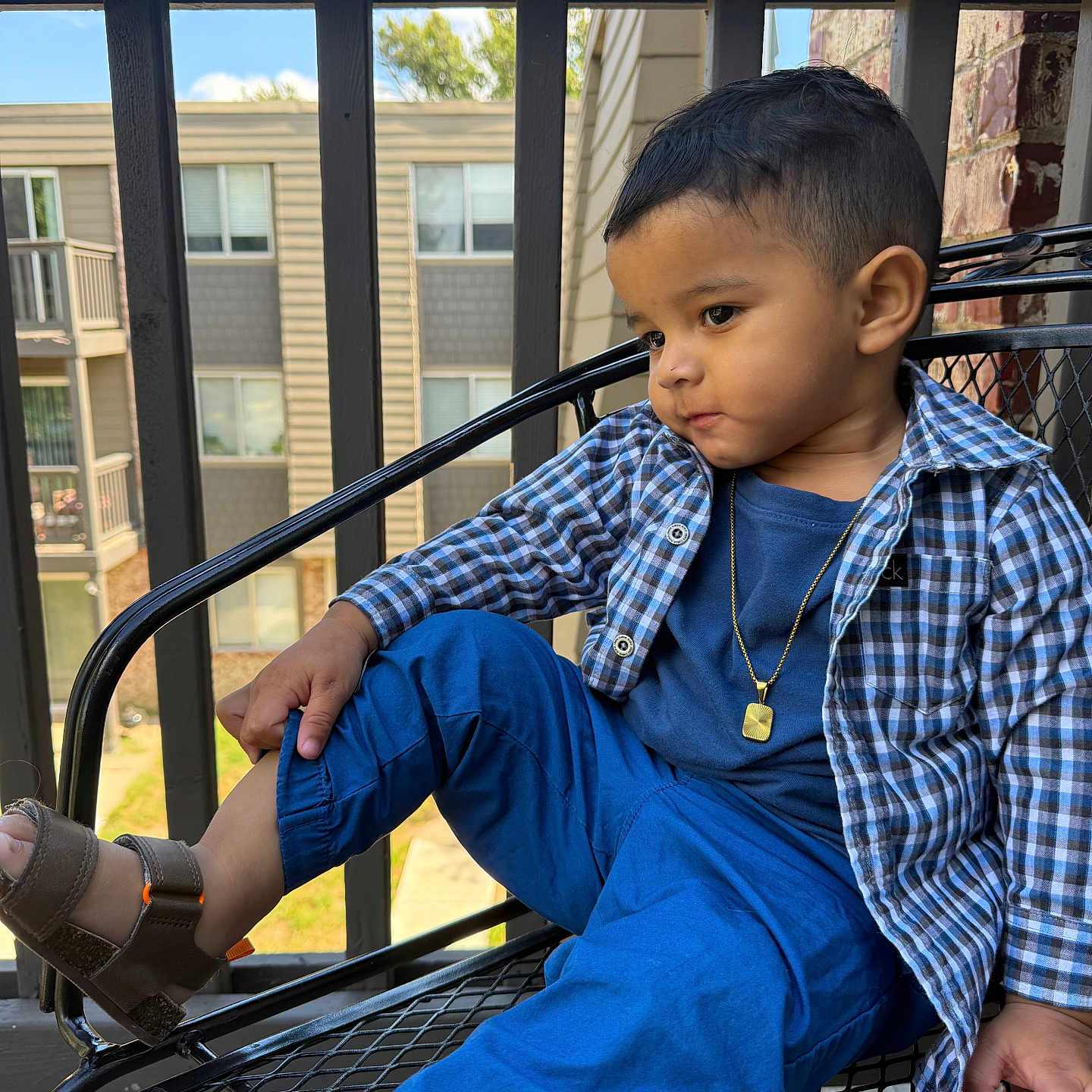 Liam Herrera Zarraga joined the competition — help win amazing prizes! child, toddler, boy, portrait, sitting, chair, outdoor, balcony, patio, blue_clothing, plaid_shirt, pants, sandals, necklace, short_hair, looking_away, face, apartment_building, railing, daylight