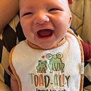 Deluca is registered to the contest to win money with this photo: baby, smile, bib, frog, embroidery, hand, infant, cute, clothing, red_shirt, joy, happy, closeup, face, mouth_open, seated, fabric, text, love, family
