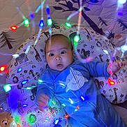 Abdiel joined the competition — help win amazing prizes! baby, infant, onesie, blue_clothing, face, cushion, animal_print, string_lights, colorful_lights, glowing, indoors, playmat, patterned, shark_design, cute, child, newborn, expression, lying_down, soft_toys