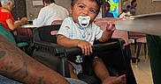 Rashaad joined the competition — help win amazing prizes! baby, casual_clothing, chair, child, curly_hair, cute, decor, dining_room, family, flower_tree, high_chair, indoor, pacifier, people, person, pink_flowers, portrait, restaurant, table, toddler