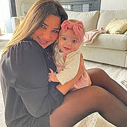 Clara participe au concours pour gagner de l'argent avec cette photo : woman, baby, smiling, indoor, living_room, couch, cushion, pink_bow, black_dress, tights, happy, hugging, child, person, floor, carpet, window, natural_light, casual, portrait