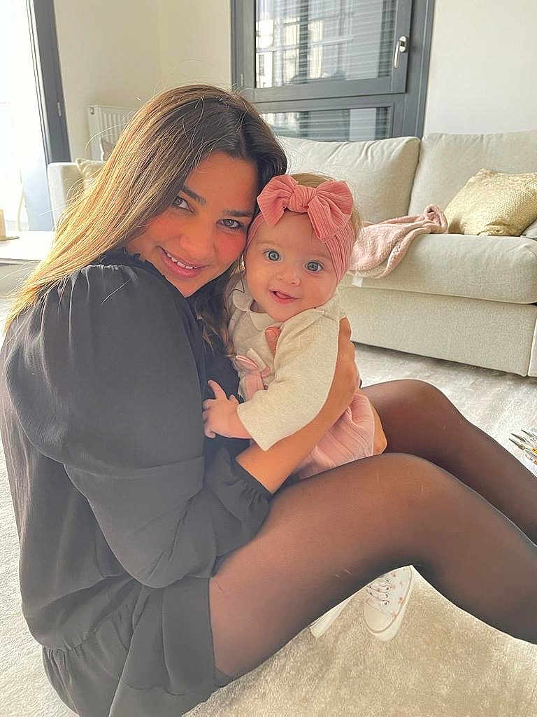 Clara participe au concours pour gagner de l'argent avec cette photo : woman, baby, smiling, indoor, living_room, couch, cushion, pink_bow, black_dress, tights, happy, hugging, child, person, floor, carpet, window, natural_light, casual, portrait