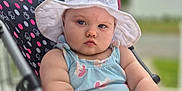 Vanessa is registered to the contest to win money with this photo: baby, child, stroller, white_hat, blue_outfit, outdoor, serious_expression, polka_dot, seat, arm, face, person, summer, sunhat, infant, cute, portrait, background_blur, daytime, baby_clothing