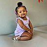 toddler, child, smiling, sitting, purple_outfit, beads, hair, sandals, couch, indoor, cute, happy, girl, portrait, fashion, skin, face, young_child, comfort, seated