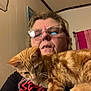 woman, glasses, cat, orange_tabby, indoor, selfie, shirt, towel, wall, curious, pet, feline, face, person, home, casual, closeup, portrait, animal, resting
