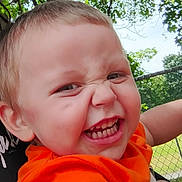 Junior is registered to the contest to win money with this photo: child, toddler, face, expression, orange_shirt, outdoor, tree, green_leaves, fence, grass, playful, close_up, person, smile, cute, happy, nature, daylight, skin, hair