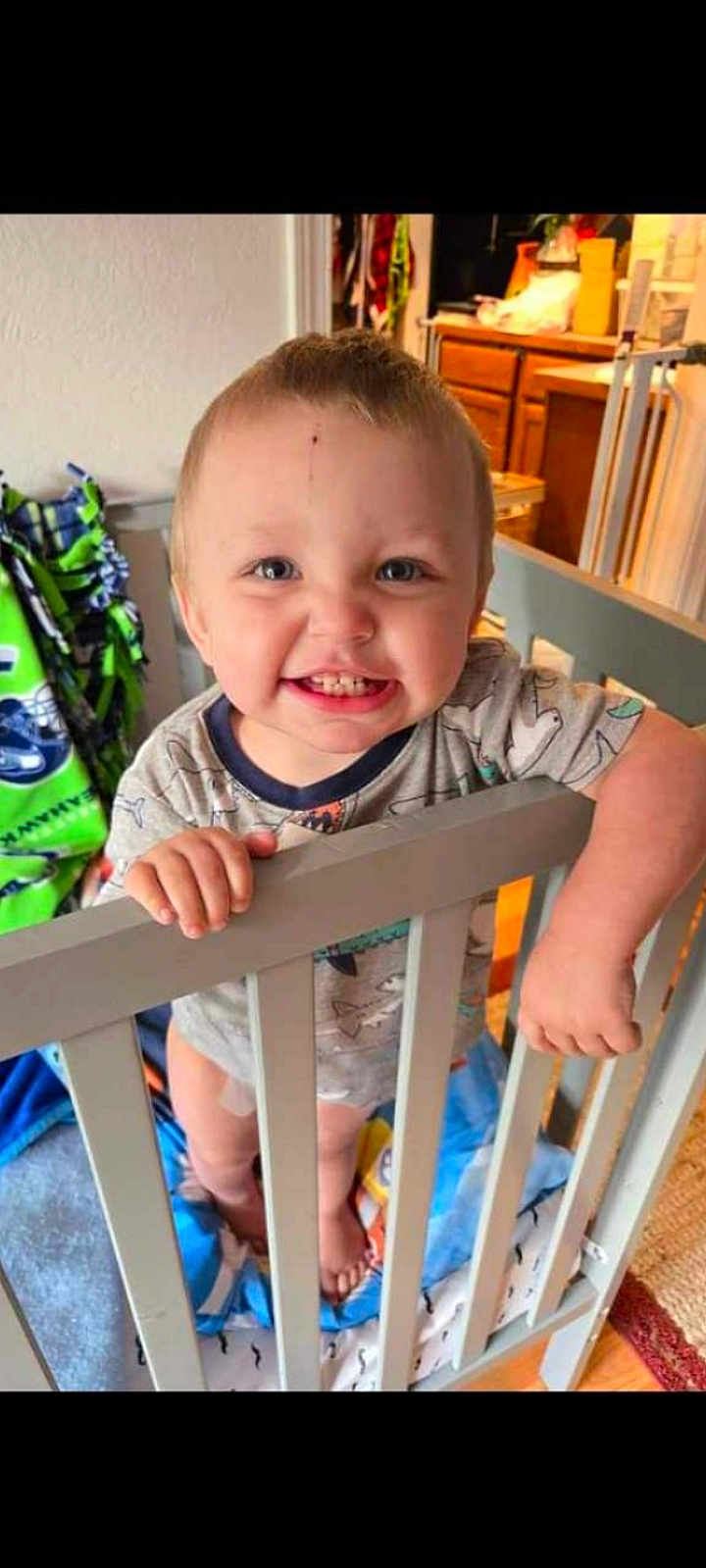 Junior is registered to the contest to win money with this photo: toddler, crib, baby, smile, happy, indoors, clothing, blanket, hand, face, foot, wooden_furniture, home, child, room, person, blue, gray, cute, portrait