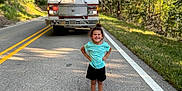 Gracelynn is registered to the contest to win money with this photo: child, girl, smiling, road, fire_truck, vehicle, trees, greenery, sunlight, outdoor, nature, asphalt, sidewalk, grass, summer, casual_clothing, sandals, happy, portrait, standing