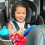 Gracelynn is registered to the contest to win money with this photo: child, car_seat, seatbelt, tablet, blanket, curly_hair, smiling, car_interior, window, drink_cup, sippy_cup, red_blanket, gray_shirt, happy, young_child, seat, automobile, safe, casual_clothing, daylight