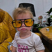 Mia a rejoint le concours — aidez-le/la à gagner de superbes lots ! toddler, child, sunglasses, yellow_glasses, oversized_glasses, spiky_hair, white_shirt, printed_shirt, leather_chair, mustard_chair, wooden_table, indoor, lamp, potted_plants, flower, portrait, serious_expression, hand, sitting, cute