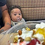 baby, child, food, dessert, whipped_cream, fruit, table, bib, tattoo, arm, patterned_background, curly_hair, wide_eyes, indoor, seat, person, closeup, expression, curious, blurred_foreground