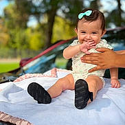 Samantha is registered to the contest to win money with this photo: baby, child, infant, smiling, happy, outdoor, portrait, sitting, blanket, bow, hair, face, hand, adult_hand, onesie, shoes, car, trees, sunlight, family