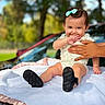 baby, child, infant, smiling, happy, outdoor, portrait, sitting, blanket, bow, hair, face, hand, adult_hand, onesie, shoes, car, trees, sunlight, family