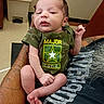 newborn, baby, infant, child, portrait, face, camouflage, shirt, star, text_on_clothing, held, adult, arm, hand, sitting, indoor, hospital, trash_can, yellow, funny