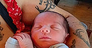 Orion is registered to the contest to win money with this photo: newborn, baby, sleeping, infant, portrait, close_up, face, hands, blanket, red_blanket, blue_clothing, adult_arms, tattoos, cuddling, indoors, wood_floor, playpen, graco, peaceful, family