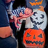 adult, arms, baby, beard, caregiving, cradling, dark_background, decoration, family, festive, halloween, indoor, infant, jack_o_lantern, newborn, orange, portrait, pumpkin, sleeping, striped_clothing