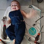 Paul participe au concours pour gagner de l'argent avec cette photo : arm_movement, baby, blue_clothing, child, denim_overalls, face, floor_tiles, hand_tools, happy, indoor, infant, lying_down, motion_blur, person, playful, screwdriver, smile, tools, towel, wrench