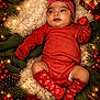 baby, blanket, christmas_lights, cinnamon_sticks, cozy, festive, gift_box, green_knitwear, headband, holiday_decor, indoors, infant, pine_cones, portrait, red_berries, red_clothing, smiling, socks, star_anise, wooden_floor