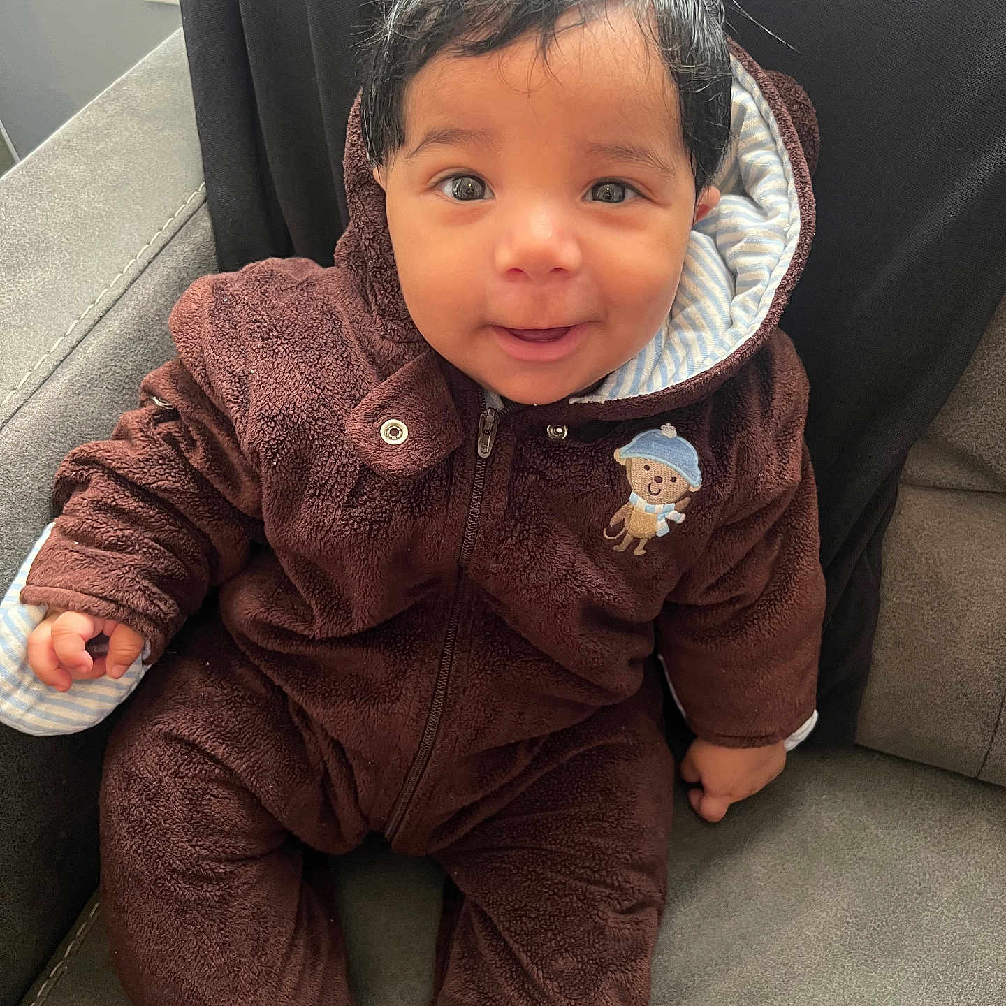 Luis Padilla is registered to the contest to win money with this photo: baby, brown_clothing, child, clothing, cozy, cute, expression, face, furniture, hair, happy, hood, indoor, monkey_slippers, onesie, person, portrait, seated, smiling, toddler