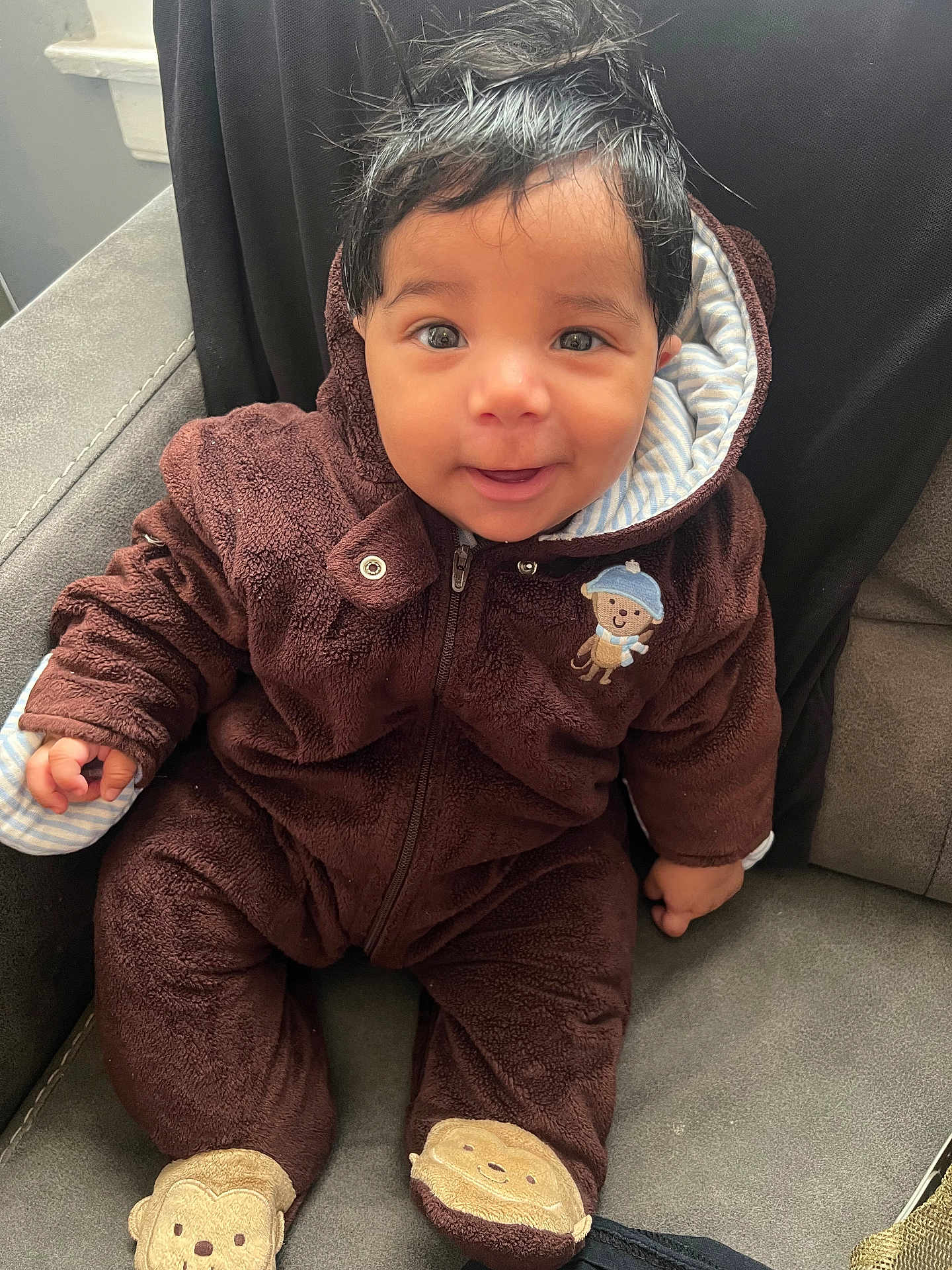 Luis Padilla is registered to the contest to win money with this photo: baby, brown_clothing, child, clothing, cozy, cute, expression, face, furniture, hair, happy, hood, indoor, monkey_slippers, onesie, person, portrait, seated, smiling, toddler