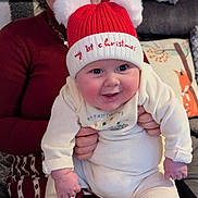 Raphael is registered to the contest to win money with this photo: baby, child, adult, smile, hat, red_hat, christmas, holiday, indoor, couch, blanket, pillow, woman, holding, cute, warm_clothing, festive, happy, face, portrait
