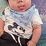 baby, child, tongue_out, blue_eyes, bib, clothing, mickey_mouse, tshirt, shorts, sitting, hand, cute, playful, indoor, portrait, infant, expression, patterned_background, young_child, smiling