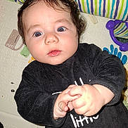 Maé a rejoint le concours — aidez-le/la à gagner de superbes lots ! baby, child, infant, curly_hair, black_shirt, text_on_clothing, hands, face, eyes, pillow, striped_pillow, play_mat, colorful, indoor, closeup, portrait, expression, person, cute, looking_up