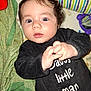 baby, infant, child, person, face, hands, clothing, black_outfit, text_on_clothing, blanket, bedding, toy, indoor, curly_hair, blue_eyes, mustache_graphic, lying_down, close_up, expression, cute