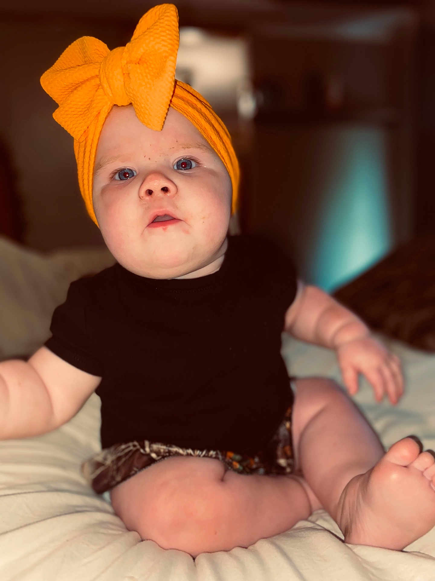 Oakley joined the competition — help win amazing prizes! baby, child, headwrap, orange, bow, black_shirt, shorts, barefoot, sitting, curious, indoor, soft_lighting, chubby, wide_eyes, cute, infant, pale_skin, portrait, closeup, bed