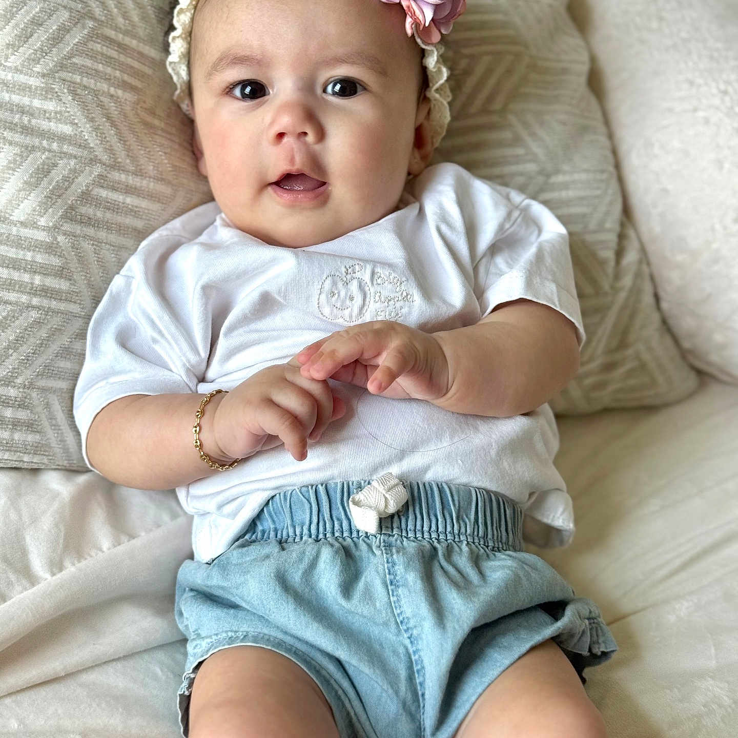 Aria is registered to the contest to win money with this photo: baby, child, infant, headband, flower, cushion, white_shirt, denim_shorts, bracelet, indoor, portrait, cute, smiling, person, skin, hand, legs, seated, cozy, soft_fabric