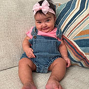 Iliany Diaz joined the competition — help win amazing prizes! baby, child, infant, smile, denim_overalls, pink_bow, headband, couch, pillow, striped_pillow, sitting, indoor, cute, happy, portrait, person, skin, hand, foot, expression