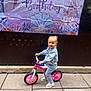 child, toddler, smile, balance_bike, toy_bike, pink_wheels, denim_jacket, jeans, sneakers, birthday_banner, party_decoration, seashell_graphic, castle_graphic, outdoor, paved_ground, wall, happy, portrait, playful, cute