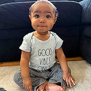 Novarae is registered to the contest to win money with this photo: baby, child, clothing, couch, cross_legged, curious, cute, floor, furniture, indoor, infant, natural_light, person, portrait, rug, sitting, smiling, striped_pants, text_on_clothing, tshirt