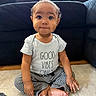 baby, child, clothing, couch, cross_legged, curious, cute, floor, furniture, indoor, infant, natural_light, person, portrait, rug, sitting, smiling, striped_pants, text_on_clothing, tshirt