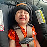 child, toddler, car_seat, smiling, happy, beanie, orange_shirt, seatbelt, car_interior, safety, cute, baby, face, headwear, portrait, transportation, travel, young_child, sitting, daylight