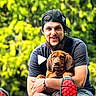 man, dog, puppy, outdoor, smiling, beard, cap, casual_clothing, greenery, nature, happy, sitting, hugging, pet, red_shoes, shorts, sunlight, leisure, portrait, friendship