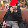 person, kitten, cat, indoor, couch, black_sweater, jeans, plant, shopping_center, table, phone, bracelet, jacket, casual, seated, holding, cute, fluffy, relaxed, public_space