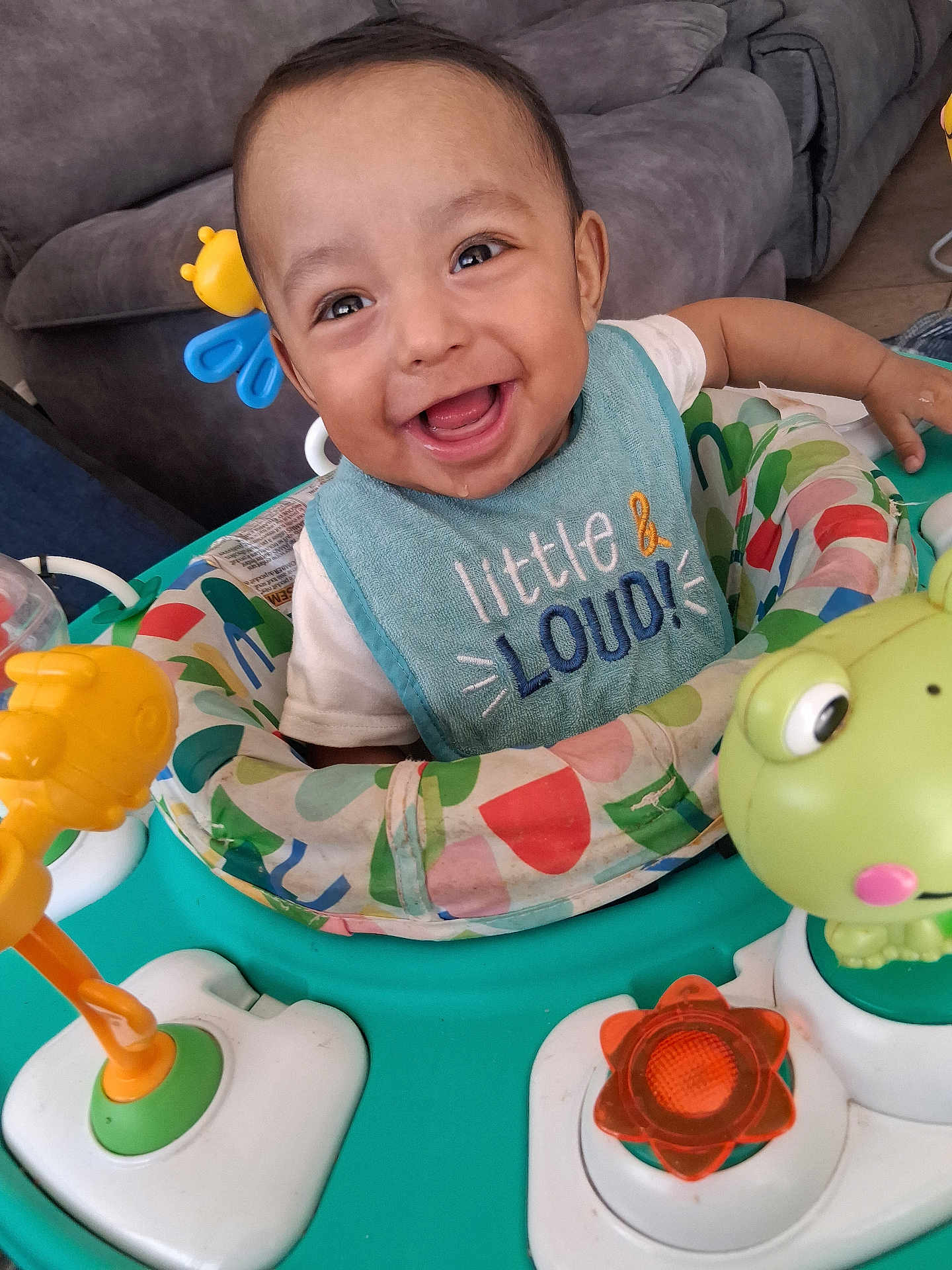 Osiris is registered to the contest to win money with this photo: baby, smiling, bib, toy, activity_center, indoor, child, happy, face, furniture, couch, colorful, playful, infant, person, clothing, cute, cheerful, closeup, portrait