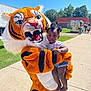 toddler, child, costume, tiger, outdoor, sunny, person, holding, grass, sidewalk, food_truck, people, building, trees, blue_sky, daylight, cute, festival, event, fun
