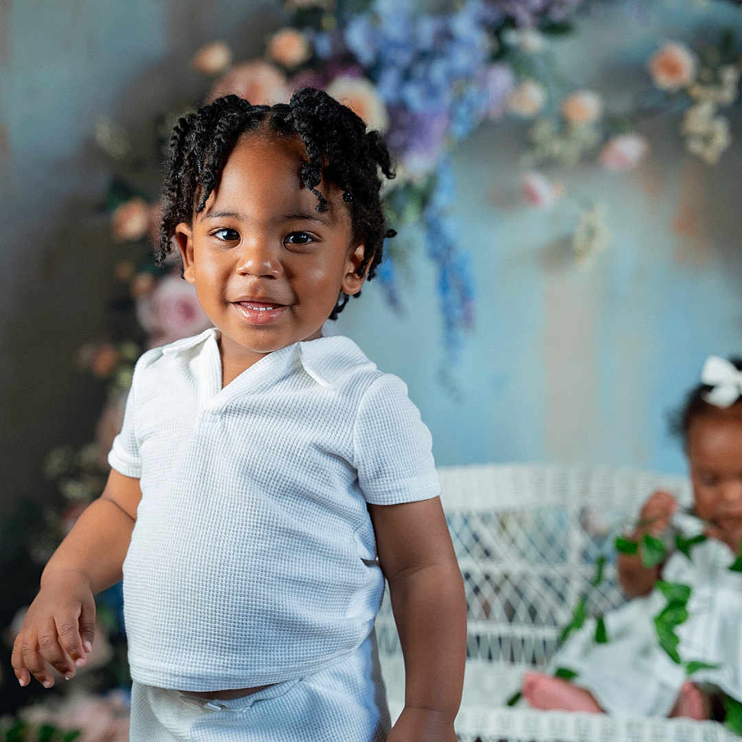 Graham is registered to the contest to win money with this photo: baby, black_hair, child, curly_hair, cute, decor, floral_backdrop, flowers, greenery, happy, indoor, portrait, shirt, shorts, smiling, standing, toddler, two_children, white_clothing, wicker_bench