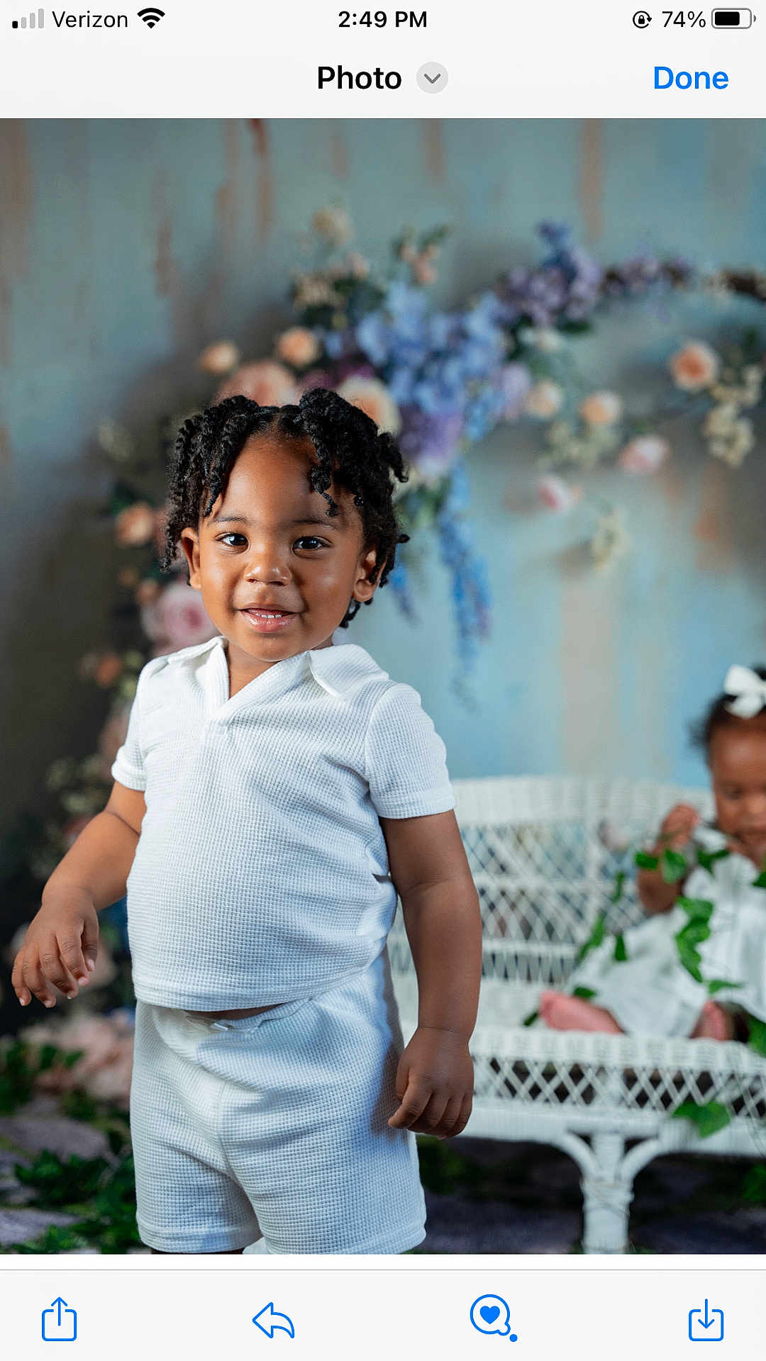 Graham is registered to the contest to win money with this photo: toddler, child, white_clothing, shorts, shirt, standing, smiling, flowers, floral_backdrop, wicker_bench, greenery, indoor, portrait, cute, black_hair, curly_hair, baby, happy, two_children, decor