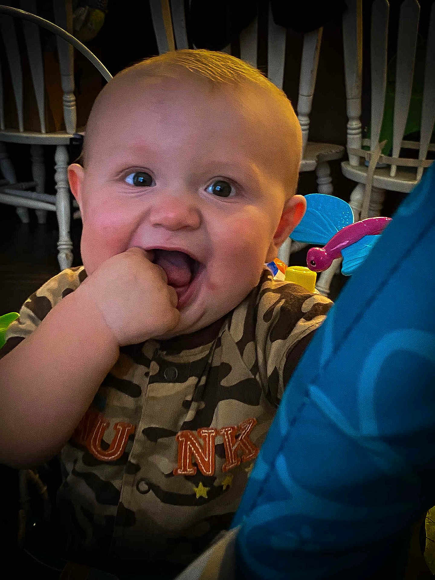 Asher is registered to the contest to win money with this photo: baby, child, smiling, camo_clothing, hand_in_mouth, indoor, furniture, chair, toy, playful, cute, portrait, happy, infant, person, face, bright_eyes, closeup, home, fun